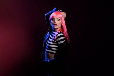 Portrait of sexy funky rocker posing in dark shadows, feeling attractive and confident in studio. Young woman with pink hair and stylish fashion, wearing biker jacket over colorful background.