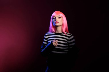 Caucasian attractive woman advertising punk fashion trend in dark shadow light, standing over pink background. Beautiful stylish person wearing leather jacket and pink hair wig in studio.