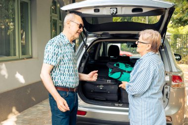 Elderly man and woman sitting in driveway with car, leaving on holiday vacation adventure after loading travelling bags and suitcases in automobile trunk. Leaving on journey with trolley for leisure.