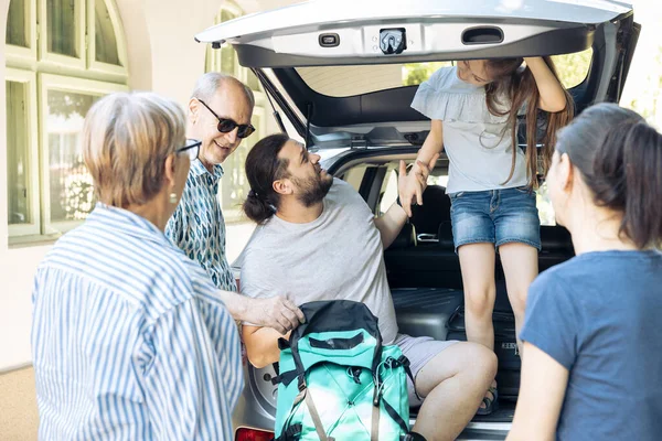 Relaxed people preparing to leave on adventure, putting travel bags and suitcase in automobile trunk. Big family with parents, grandparents and small child travelling on summer holiday trip.