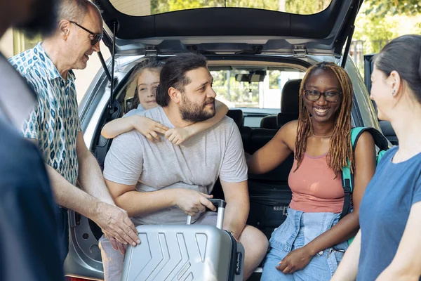 Diverse family and friends loading trolley in automobile trunk, preparing to leave on holiday trip. Travelling by car on summer vacation, going to seaside destination with multiethnic people.