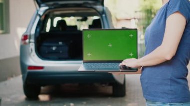 Woman having laptop with green screen chroma key background. Person with portable computer having isolated template mock up display while people putting baggage inside car trunk in background.