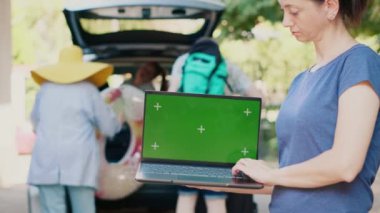 Person with modern computer having isolated template mock up screen while people putting luggage inside vehicle trunk in background. Woman having laptop with green screen chroma key background.