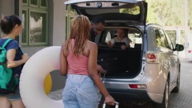 Multiethnic people loading luggage in vehicle trunk while unpatient little girl hurrying them. Mother and family friends putting voyage baggage in car while getting ready for field trip departure.