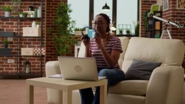 African american employee working remotely on laptop, listening to music on headphones and sitting on sofa. Businesswoman browsing internet for remote job at home, online network.