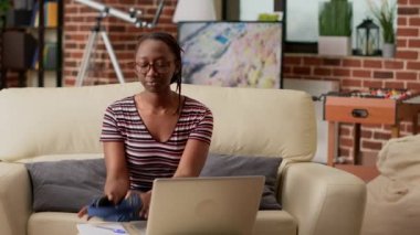 African american woman listening to music and working remotely on laptop, using internet network to do remote business job. Browsing online website and wearing headphones at home.