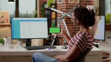 Young adult holding smartphone with horizontal greenscreen at home office desk. Looking at blank copyspace template with mockup background and isolated chroma key display. Tripod shot.
