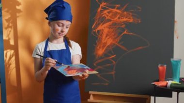 Portrait of young child using orange color paint on canvas, creating art masterpiece with paintbrush and watercolor dye. Little girl making artwork design with aquarelle palette. Handheld shot.