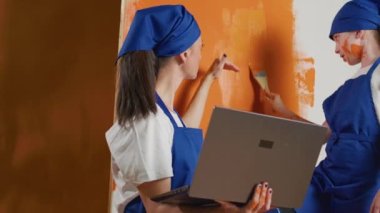Small family using orange paint to renovate apartment walls with online inspiration on laptop, painting room with paintbrush. Browsing model on computer, internet decorating. Handheld shot.
