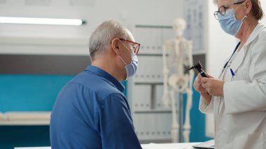 Senior physician doing ear examination with otoscope, consulting infection with otology instrument in cabinet. Otologist using medical tool at checkup visit during covid 19 pandemic.
