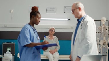 Retired woman waiting to receive sickness diagnosis from medical staff in cabinet, to cure disease with medicine. Sitting in doctors office to find exam results from diverse team of specialists.