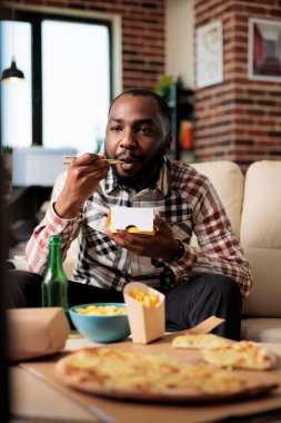 Relaxed man enjoying noodles package from takeaway delivery, eating food with chopsticks in front of television movie. Watching film on tv channel program, having fun with takeout meal.