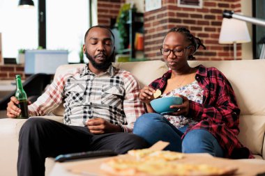 Relaxed people eating chips from bowl and drinking beer, having fun together with fast food takeaway meal. Enioying takeout delivery and watching movie or film on television, entertainment.