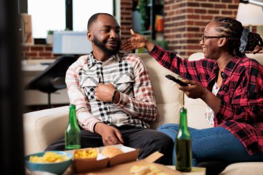 Young partners laughing and watching television together, eating fast food delivery meal while girlfriend switches channels with tv remote control. Takeaway food and beer to have fun with film.
