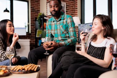 Diverse group of people having wine party at home to celebrate friendship anniversary. Smiling african american man sitting on sofa while celebrating job promotion with best friends.