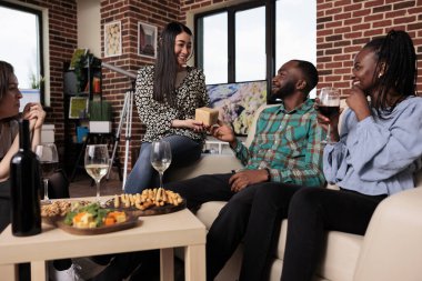 Smiling diverse people enjoying wine party at home while exchanging presents and drinking wine. Young multiethnic group of friends bonding while giving each other gifts at home in living room.