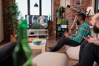 Multiethnic group of friends playing shooting video games on tv console, drinking beer at social gathering with people. Having fun with gaming competition and leisure activity at house party.