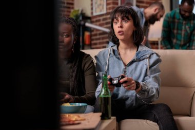 Multiethnic friends using tv console to play video games at home, gathering with people at house party. Happy women playing competition on television, relaxing together with beer bottles.