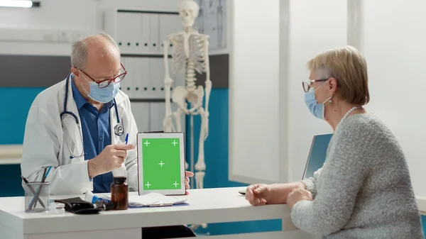 Physician and patient with face mask looking at greenscreen on digital tablet at appointment. Mockup blank template with isolated chroma key background and copyspace on gadget.