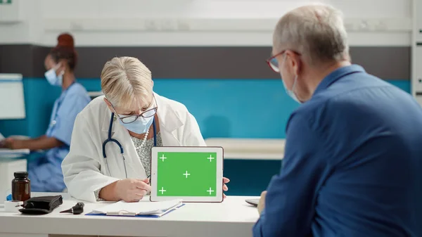 Doctor and retired person looking at greenscreen on tablet during covid 19 pandemic in medical cabinet. Blank mockup background with isolated copyspace and chroma key template.