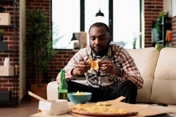 Relaxed person eating slice of pizza in front of television at home, having takeaway delivery meal with bottles of beer. Watching film on television and enjoying fast food takeout to eat dinner.