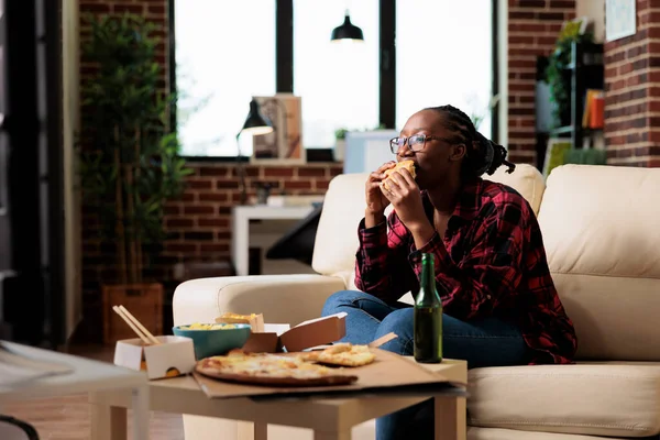 African american woman eating burger from fast food delivery takeout, watching movie on tv channel program. Serving takeaway meal to enjoy leisure activity with film on television.