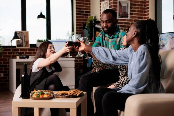 Diverse people celebrating friendship event while having wine party at home. Multiethnic group of cheerful friends clinking wine glasses while toasting and drinking alcoholic beverages.