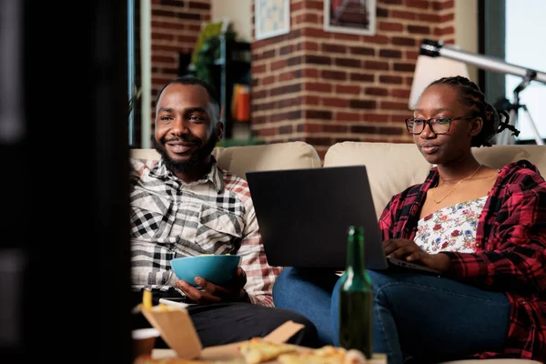 Life partners watching movie on tv and laptop to browse internet, eating fast food takeaway delivery at home. Using computer in front of television, eat pizza and chips with beer bottles.