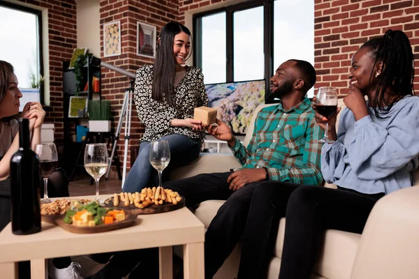 Smiling diverse people enjoying wine party at home while exchanging presents and drinking wine. Young multiethnic group of friends bonding while giving each other gifts at home in living room.
