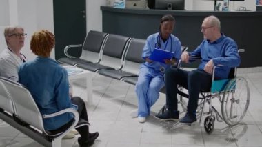 Medical assistant consulting old man with impairment, using checkup report papers. Patient with physical disability in wheelchair doing consultation with nurse in clinic waiting area.
