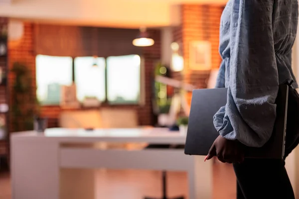 Female remote worker holding laptop in home office with modern interior and big windows on background. African american businesswoman carrying portable computer, side view