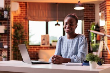 Female remote worker sitting at workplace and looking at camera in modern office. Successful freelancer working on laptop in modern corporate workspace with warm sunset light