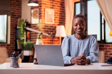 Female smiling employee working on laptop in modern home office. Young successful african american freelancer sitting at desk in room with modern interior and looking at camera