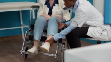 Nurse and doctor consulting woman with chronic impairment, doing physiotherapy examination in cabinet. Senior adult wheelchair user receiving alternative medicine consultation.