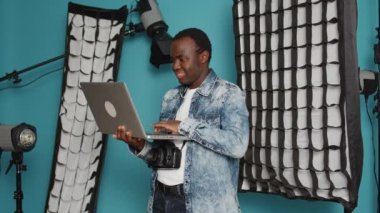 Young adult browsing internet on laptop to find photography inspiration, using professional studio shooting and lighting equipment. Looking on online website and sitting in studio backstage.