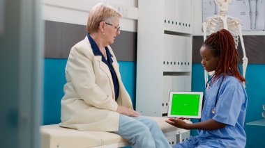 Nurse and senior patient looking at greenscreen on digital tablet in medical cabinet. Health specialist holding device with blank mockup template and isolated chromakey copyspace.