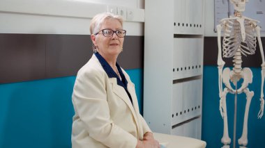 Portrait of elderly patient with disease sitting in doctor office, waiting to attend checkup examination with medical specialist at clinic. Old woman with consultation appointment in cabinet.