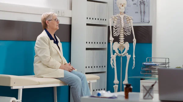 Elderly patient waiting in doctor office to attend checkup visit, having consultation appointment with physician at healthcare clinic. Old woman with disease sitting in cabinet for examination.