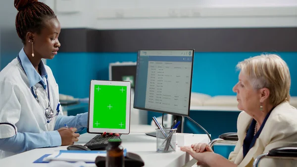 Health specialist showing greenscreen on tablet to woman with disability in cabinet. Looking at blank mockup template with isolated display and chroma key copyspace, wheelchair user.