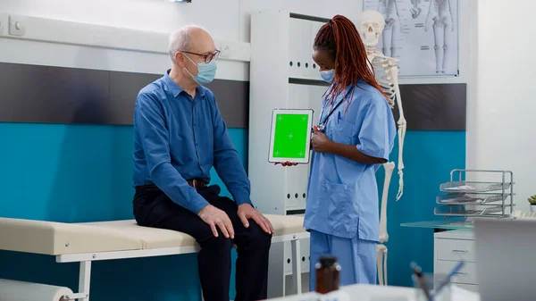 Woman and elderly man with face mask using greenscreen on tablet, isolated display. Nurse and patient looking at chromakey template with mockup background and blank copyspace in cabinet.