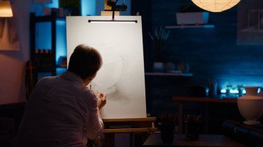 Yaratıcı yetenekleri olan yaşlı bir kadın vazo ilhamı ve sanat malzemeleriyle profesyonel sanat eserleri çiziyor. Beyaz tuval üzerinde sanat şaheseri yaratmak için araçlar ve kalemler kullanarak, çizim yapın.