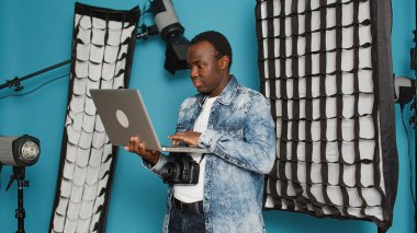 Young adult browsing internet on laptop to find photography inspiration, using professional studio shooting and lighting equipment. Looking on online website and sitting in studio backstage.