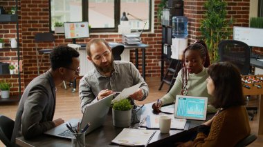Diverse team of coworkers analyzing business charts on documents, working on presentation report in boardroom. Using data on documents to plan research in corporate management meeting.