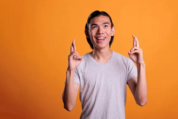 Young asian man crossing fingers, praying for luck, looking upwards. Smiling superstitious teenager getting lucky, pleading for success, making wish with hopeful facial expression