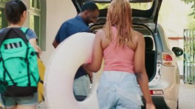 Multiethnic people loading car trunk with voyage luggage while little girl hurrying them. Diverse couple with mother and daughter putting baggage inside vehicle while going on holiday summer citybreak