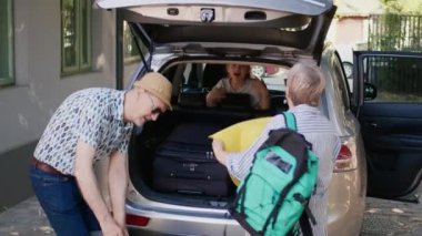Senior couple and niece going on summer field trip while putting voyage baggage and trolleys in vehicle. Grandparents loading car trunk with luggage while unpatient granddaughter hurrying them.