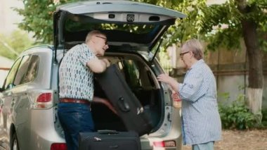 Lovely senior couple putting luggage in car trunk while getting ready for summer citybreak departure. Old people loading voyage baggage and trolleys inside vehicle while going on holiday field trip.