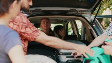 Family putting voyage baggage in car trunk while little girl hurrying them. Casual people loading vehicle with luggage and trolleys while getting ready for holiday summer citybreak departure.