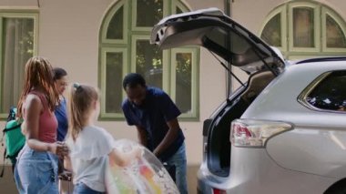 Family and african american friends going on field trip while putting baggage in vehicle. Multiethnic people going on summer citybreak while loading voyage luggage and trolleys in car trunk.