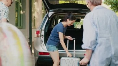 Family loading voyage luggage in car trunk while getting ready for summer holiday citybreak. Mother, daughter and grandparents putting baggage and trolleys inside vehicle while going on field trip.